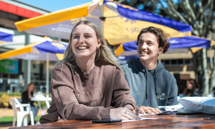 Two young people sit at an outdoor table, smiling, with umbrellas and other people in the background.