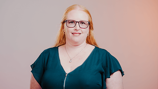 Woman with long red hair and glasses wearing a dark green top, standing against a neutral background and smiling at the camera.