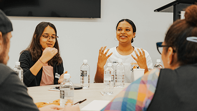 Four people sit around a table with water bottles, having a discussion; one woman gestures with her hands while speaking.