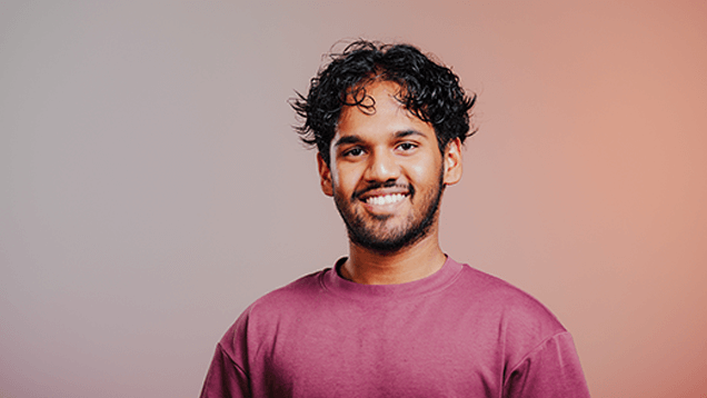 A young man with curly hair and a beard, wearing a mauve shirt, smiles while standing against a gradient pink and gray background.