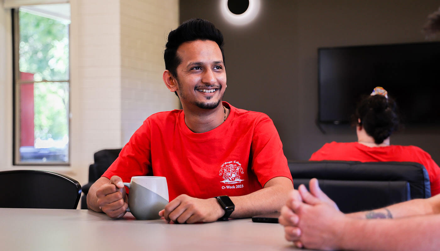 A person in a red shirt sits at a table holding a mug, smiling, with another person in the background and a TV on the wall.