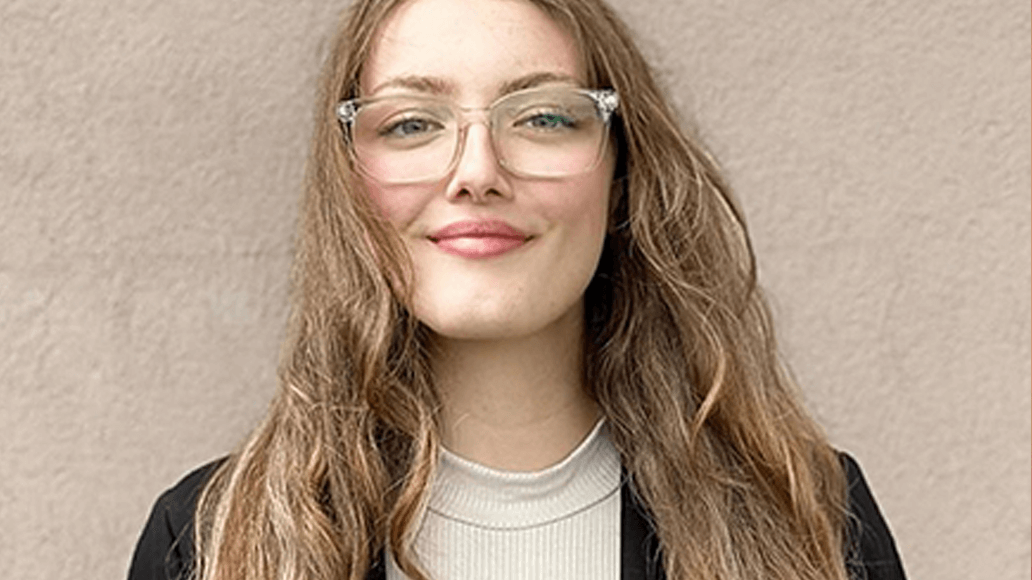 A woman with long brown hair and glasses is standing against a plain beige wall, wearing a light turtleneck and a black blazer.