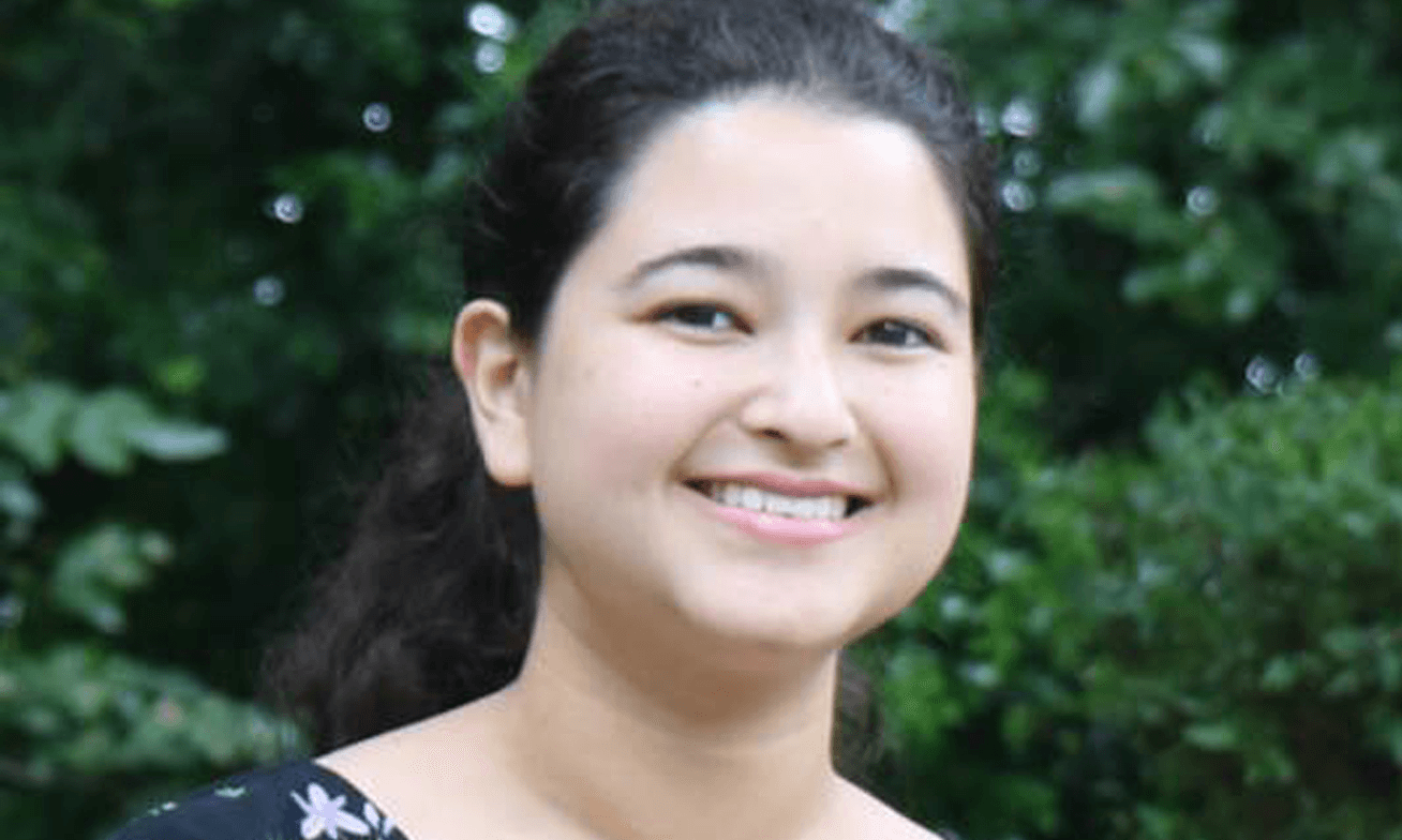 A person with dark hair pulled back smiles at the camera, standing outdoors in front of leafy green foliage.