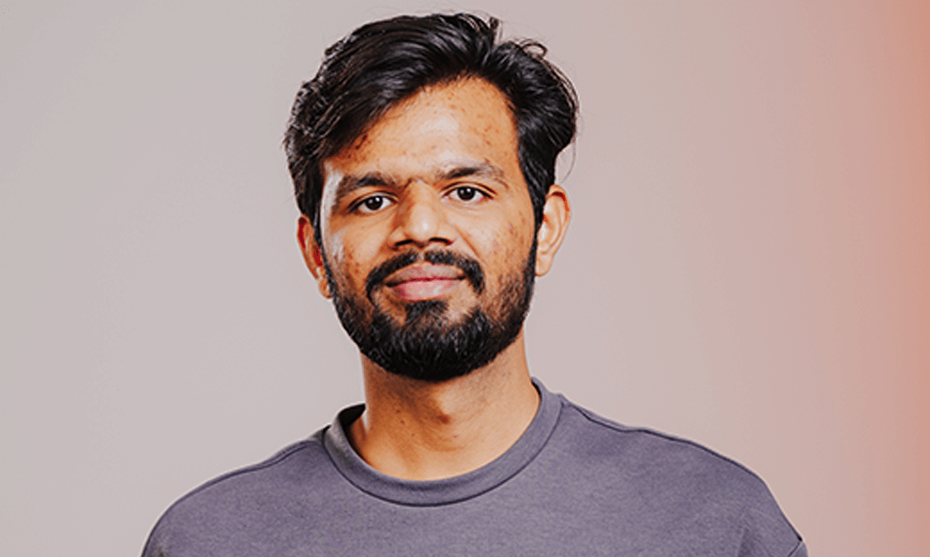 A man with a beard and mustache, wearing a gray crew-neck shirt, stands in front of a neutral gradient background.
