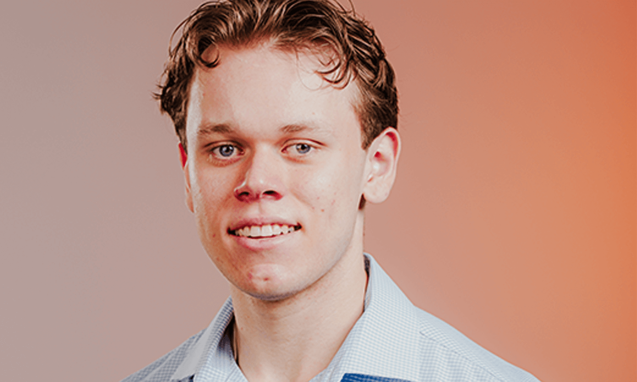 A young man with short, wavy hair wearing a light blue collared shirt, smiling slightly, posed against a gradient orange background.