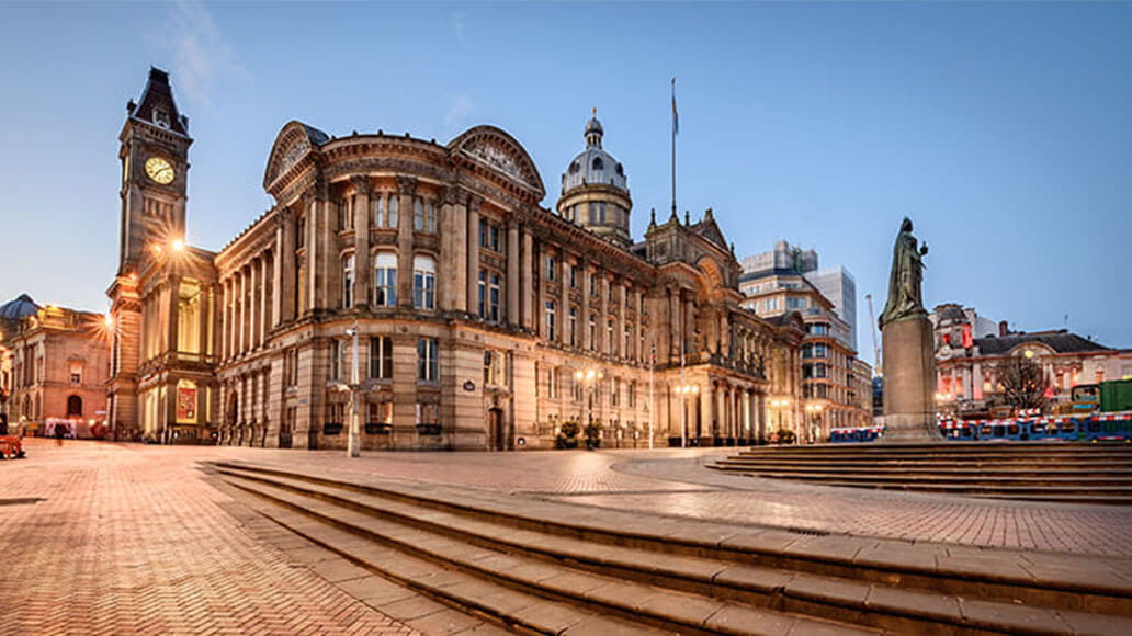 A large, historic building with columns and a clock tower stands beside a public square with steps and a statue in the foreground.