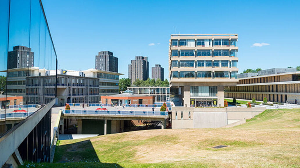 Modern university campus with concrete buildings, glass windows, and high-rise towers in the background on a clear, sunny day.