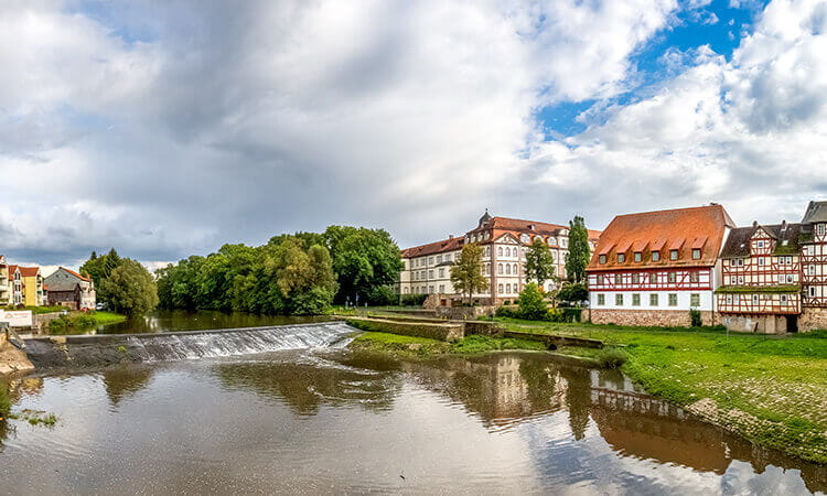 A river with a small waterfall flows past historic buildings with red roofs and timber frames, under a partly cloudy sky.