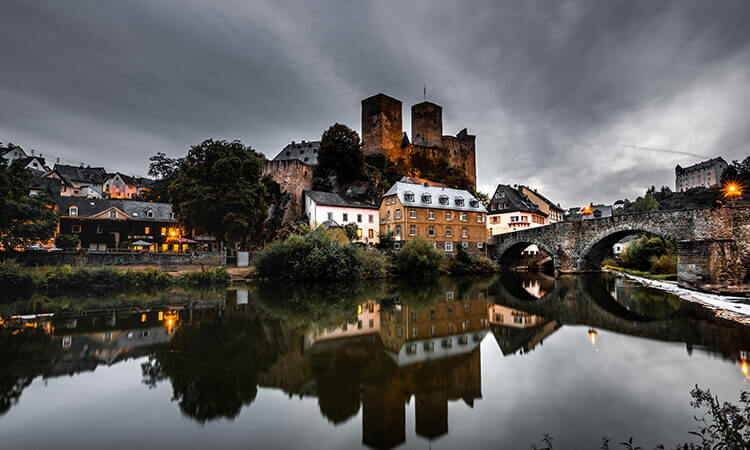 A medieval castle and stone bridge are reflected in a calm river, surrounded by old buildings and greenery under a cloudy sky at dusk.