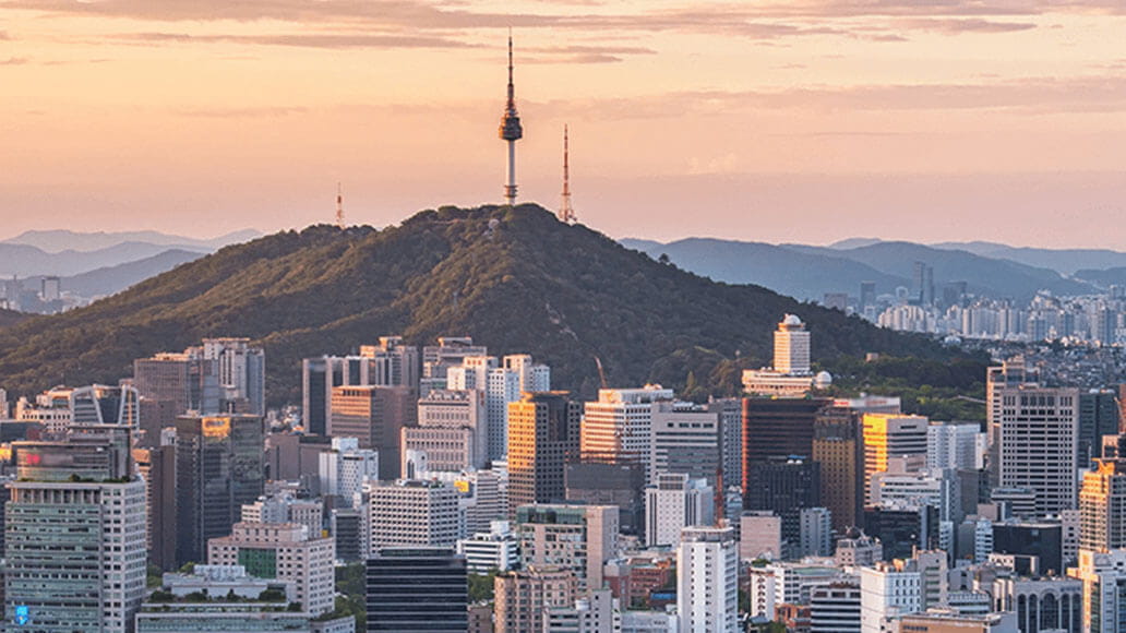 City skyline at sunset with a forested hill in the background, featuring a tall communication tower on top.