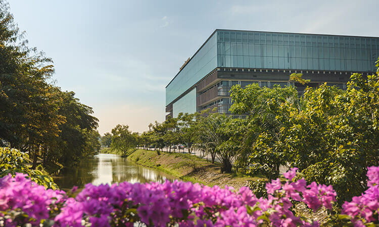 A modern glass building stands beside a canal, with green trees and pink flowers in the foreground under a clear sky.