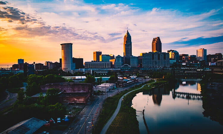 A city skyline with tall buildings is reflected in a river at sunset, under a partly cloudy sky.
