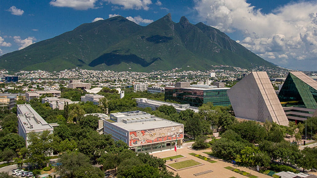 A cityscape with modern buildings and greenery is set against a backdrop of a large, distinctive mountain under a partly cloudy sky.