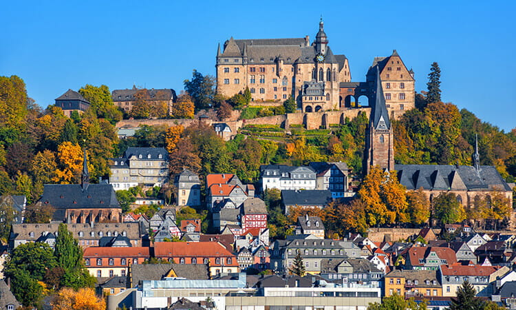 A hilltop castle overlooks a town with colorful, traditional buildings and a church, surrounded by trees with autumn foliage under a clear blue sky.