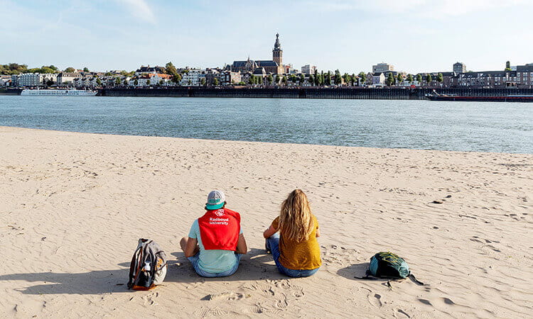 Two people with backpacks sit on a sandy riverbank facing the water, with a cityscape and church visible across the river under a clear sky.