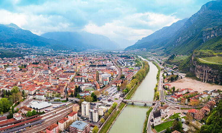Aerial view of a city with red-roofed buildings, a river flowing through the center, mountains in the background, and several bridges crossing the river.