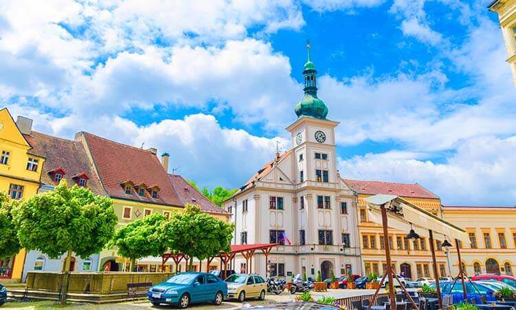 Town square with a historic white clock tower, colorful buildings, parked cars, trees, and a partly cloudy blue sky in the background.