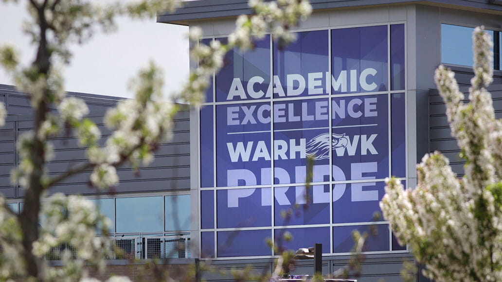 Large window display reads "Academic Excellence Warhawk Pride" on a modern building, partially obscured by blossoming tree branches in the foreground.