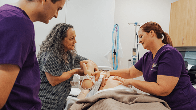 Two students practice a medical procedure on a mannequin in a clinical simulation room, they are wearing purple scrubs and are being taught by an academic in a patterned shirt.