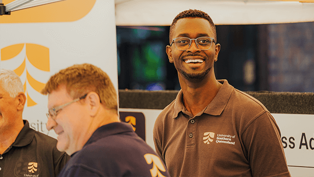 Three men wearing University of Southern Queensland shirts stand together at an event booth; one man in the center is smiling at the camera.