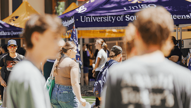 Students walk outside near tents with "University of Southern Queensland" signage during a campus event on a sunny day.