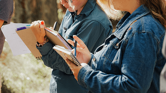 Two people in denim shirts are outdoors, holding clipboards and notebooks, writing notes.