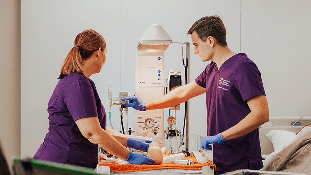 Two UniSQ Midwifery students in purple scrubs work with medical equipment next to a hospital bed, wearing blue gloves in a clinical setting.