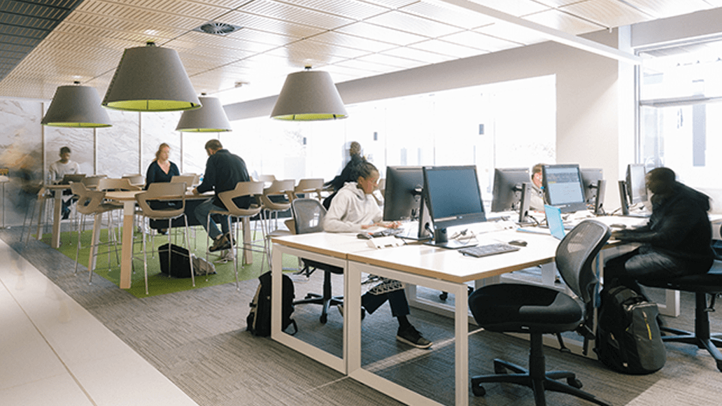 People working and studying on computers at desks and tables in a modern, well-lit library at UniSQ Springfield with large windows and hanging lights.