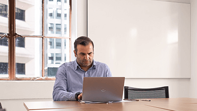 A man sits alone at a desk in an office, working on a laptop with a pen and notebook nearby. A window and blank whiteboard are in the background.
