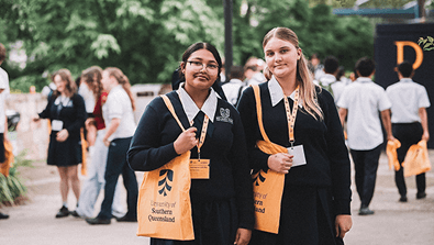 Two students in dark uniforms with University of Southern Queensland tote bags stand outdoors among a group of people.
