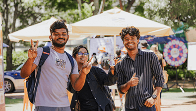 Three people stand outdoors in front of umbrellas, smiling and gesturing peace and thumbs up signs, with trees and a crowd in the background.