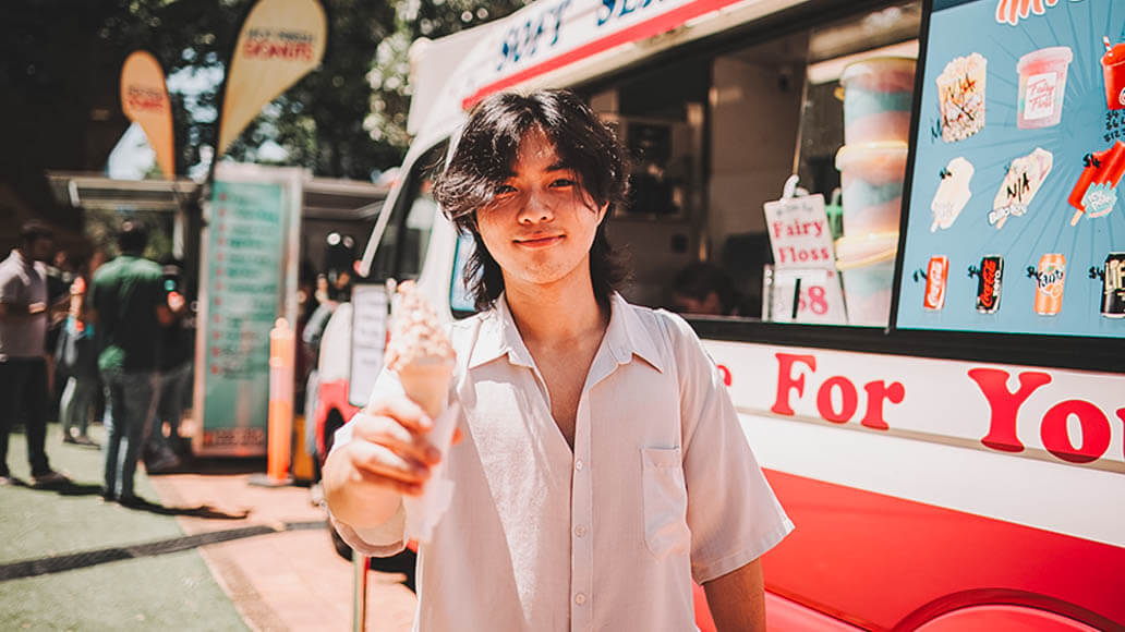 A person stands in front of an ice cream truck, holding an ice cream cone and smiling at the camera on a sunny day.