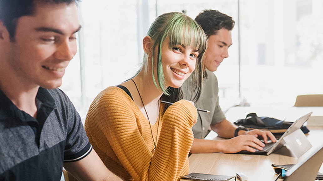 Three young adults sit at a desk working on laptops and notebooks. One person with green hair and an orange sweater smiles at the camera.