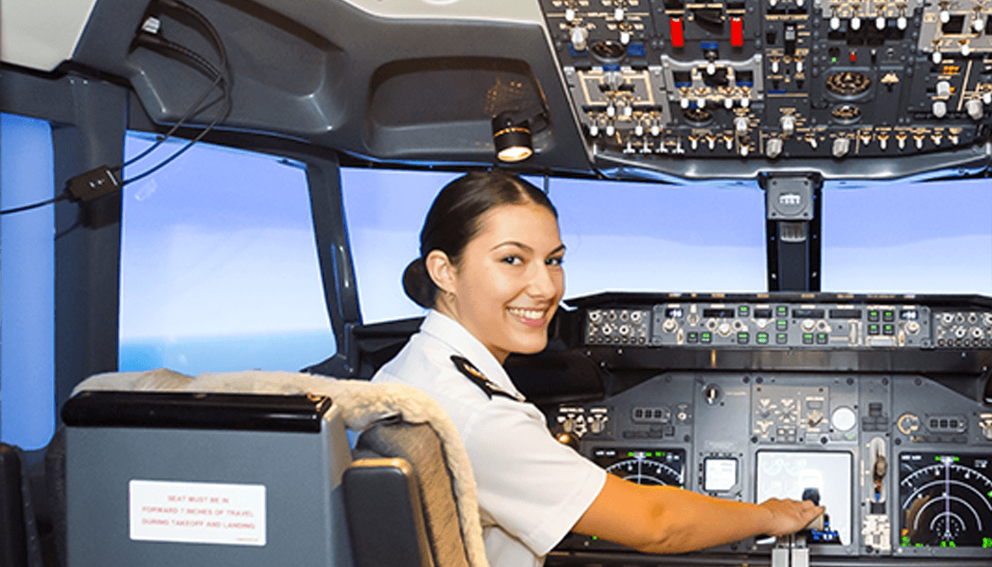 A female aviation student in uniform sits at the controls of an aircraft cockpit, smiling and looking back at the camera.