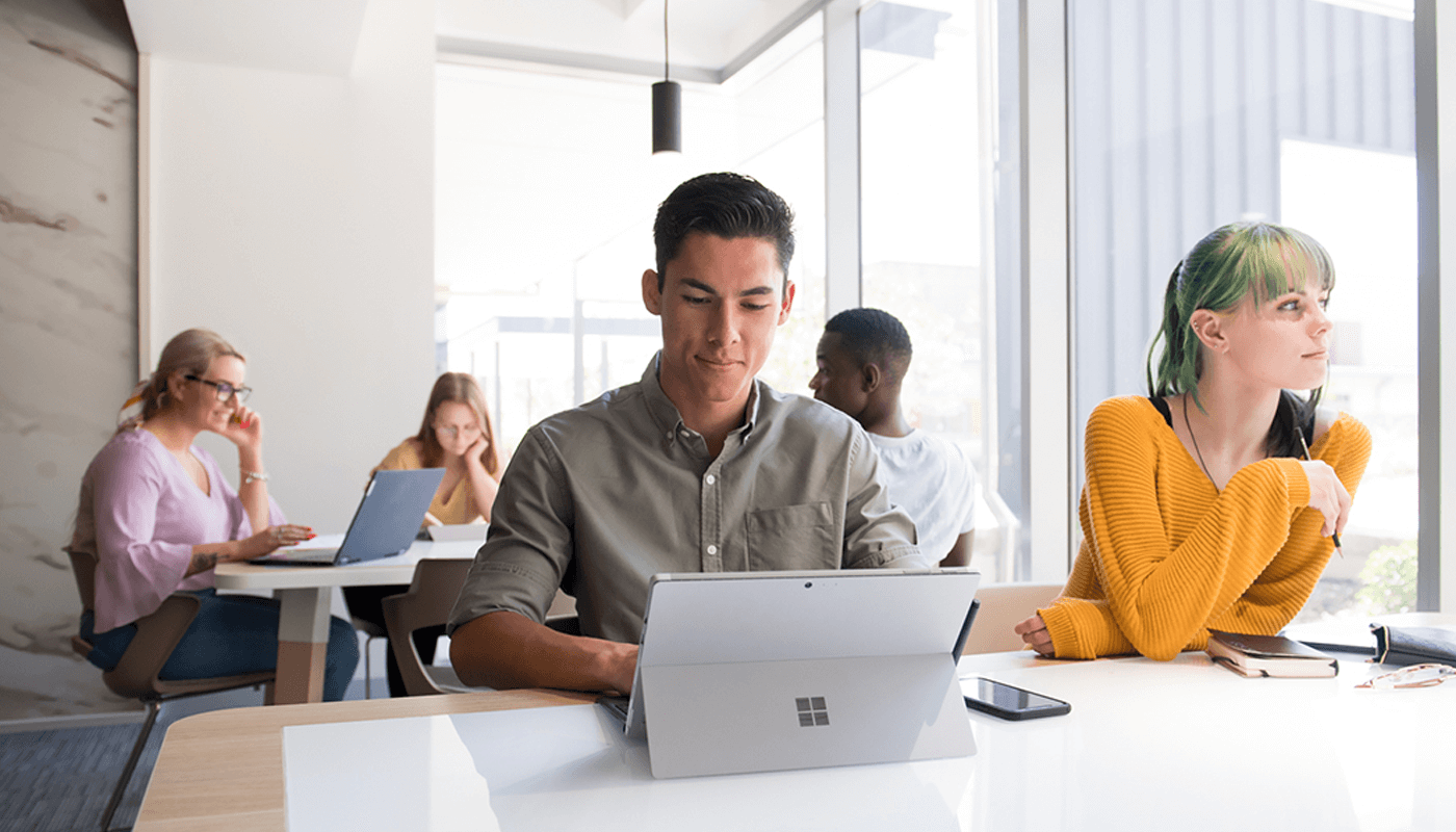 Several people work on laptops and tablets at tables in a bright, modern office or study space with large windows.