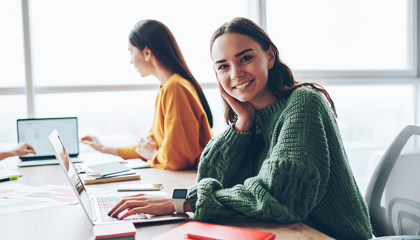 Woman in a green sweater sits at a desk with a laptop, smiling at the camera, while another person works in the background.