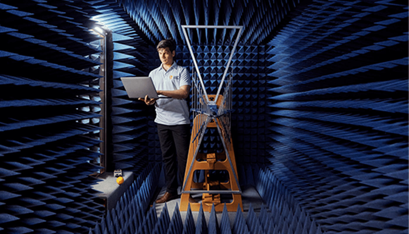 A person stands in an anechoic chamber holding a laptop near a metal antenna structure, surrounded by blue acoustic foam panels on the walls and ceiling.
