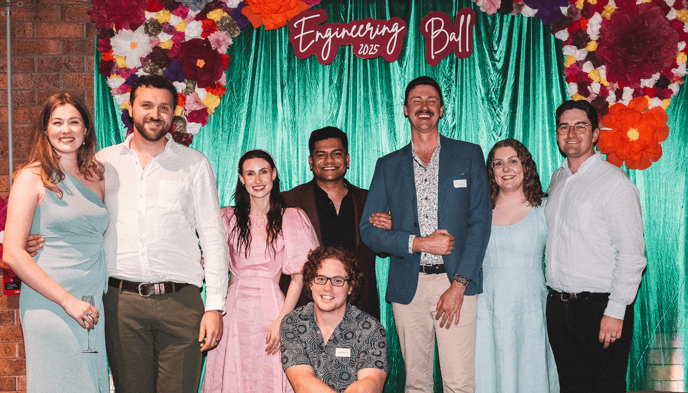 Eight people pose in front of a green curtain and colorful flower decorations at an event labeled "Engineering Ball 2025".