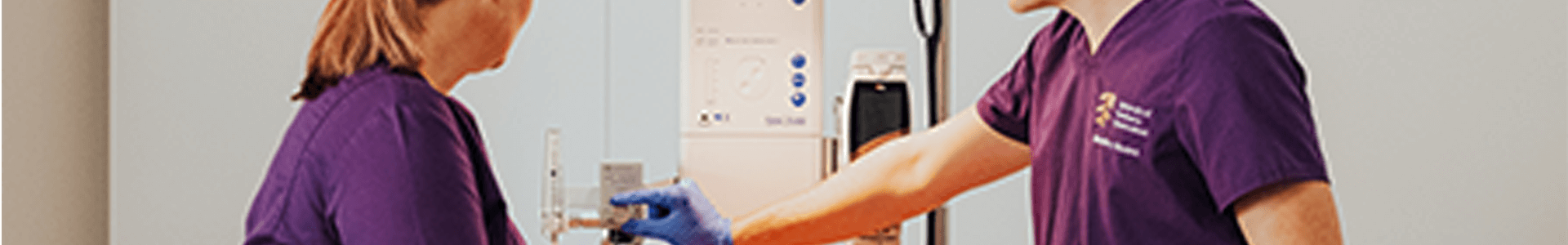 Two midwifery students in purple scrubs interact with medical equipment in a clinical setting.