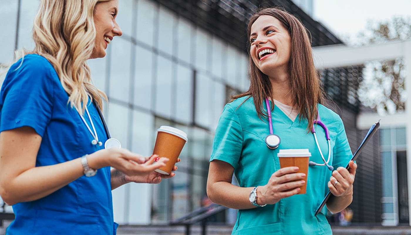 Two healthcare workers in scrubs stand outside a building, holding coffee cups, talking and smiling, with stethoscopes around their necks.