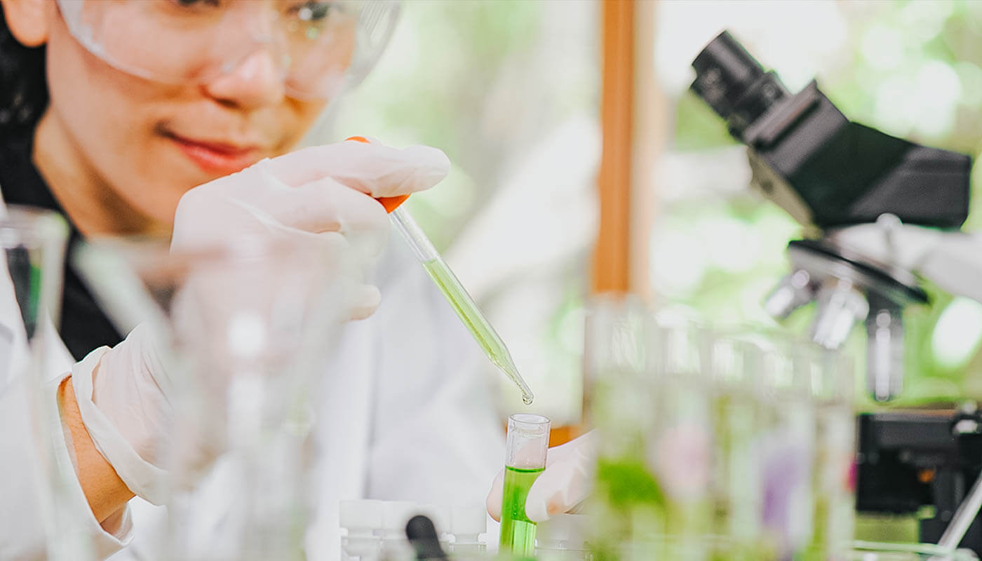 A person wearing safety goggles and gloves uses a pipette to transfer green liquid into a test tube in a laboratory, with a microscope visible in the background.
