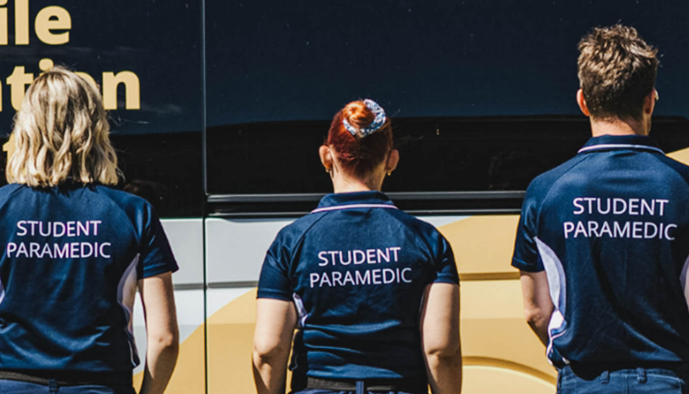 Three people wearing navy shirts labeled "STUDENT PARAMEDIC" stand with their backs to the camera in front of a dark vehicle with partial yellow text.