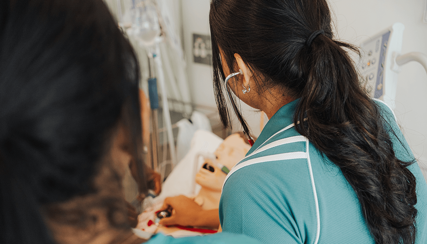 Two UniSQ nursing students in teal uniforms examine a patient using a stethoscope in a clinical setting, with medical equipment visible in the background.