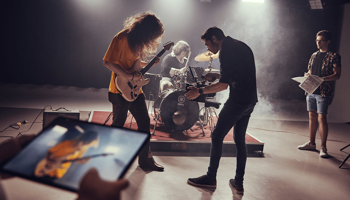 A band performs on stage with a guitarist, drummer, and two crew members; one films with a camera while another holds papers. A tablet shows the guitarist’s image in the foreground.