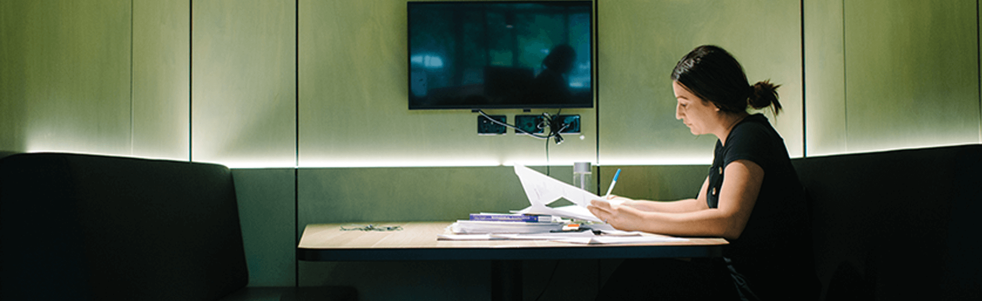 A person sits alone at a table in a modern study room, reading documents and writing, with papers and books spread out and a screen mounted on the wall.
