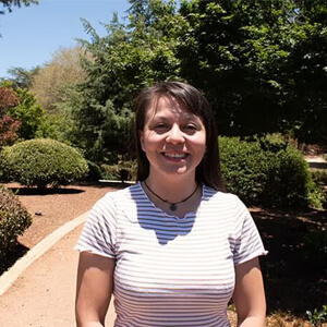 A woman in a striped shirt stands on a sunlit path in a park, surrounded by green trees and bushes.