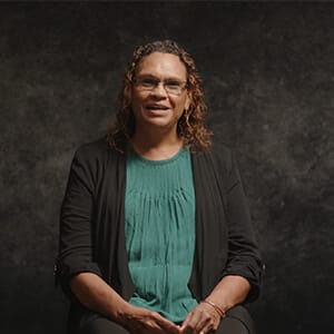 A person with curly hair, glasses, and a green top under a black jacket sits in front of a dark, textured background.