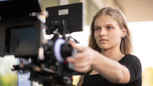 A person with long blonde hair operates a professional video camera outdoors, focusing on adjusting its lens or settings.
