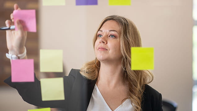 A woman writes on a pink sticky note surrounded by other colorful sticky notes attached to a glass surface.