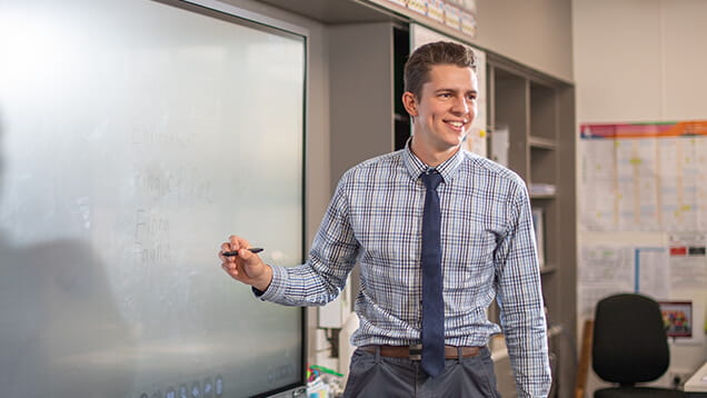 A man in a checkered shirt and tie stands by a large digital screen, holding a marker and smiling in a classroom setting.